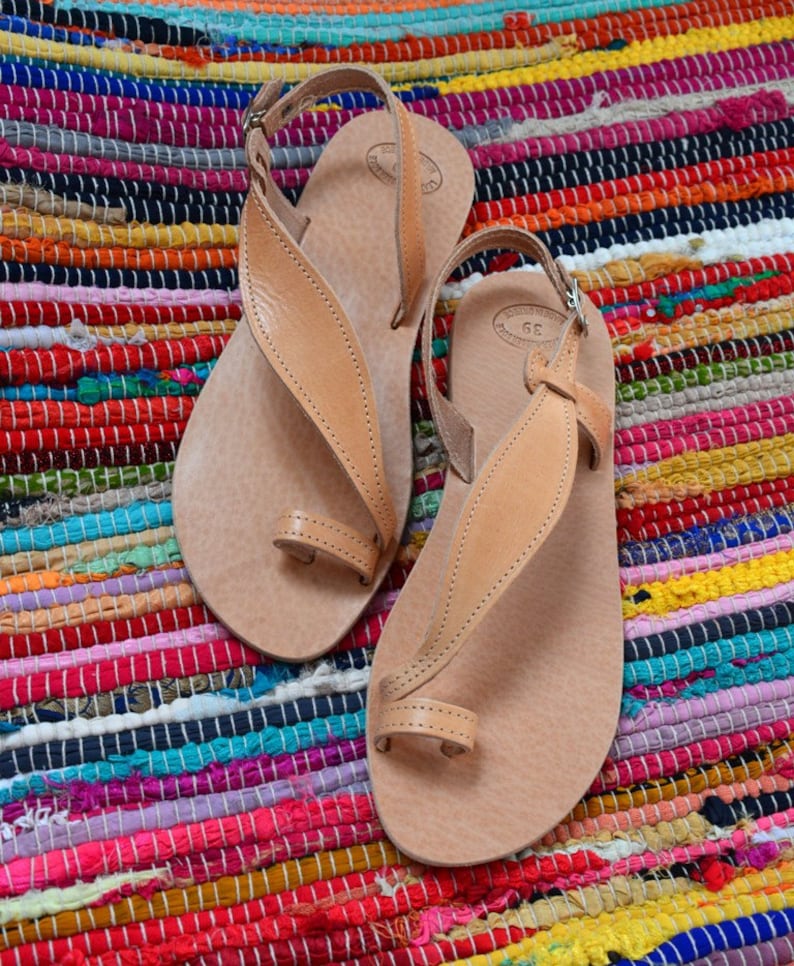 Puede incluir: Un par de sandalias de cuero marr&oacute;n claro con correas para los dedos y el tobillo. Las sandalias se exhiben sobre una colorida alfombra a rayas con tonos de rojo, rosa, azul y amarillo. Dise&ntilde;o minimalista.