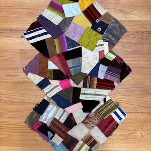 May include: A patchwork textile piece composed of numerous irregular fabric scraps in various textures and colours, including velvet, silk, and striped patterns. The assembly features a mix of deep burgundy, yellow, blue, green, and white segments.