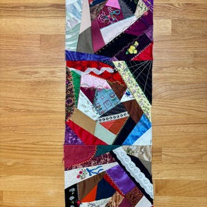May include: A rectangular crazy quilt table runner featuring a patchwork of various fabric scraps in diverse colors and textures, including satin, velvet, and lace, joined by decorative embroidery stitches and floral motifs throughout the surface.