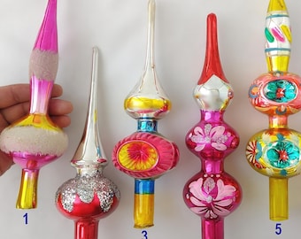 Puede incluir: Seis puntas de árbol de Navidad de vidrio vintage dispuestas en fila. Cada adorno presenta una forma de aguja alargada única con detalles pintados coloridos, acabados metálicos y acentos de purpurina. Están numerados del uno al seis.