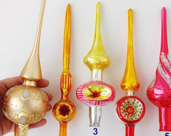 Puede incluir: Seis puntas de árbol de Navidad de vidrio dispuestas en fila. El número 1 es dorado mate con detalles de purpurina. El número 2 es naranja. El número 3 es amarillo y plateado. El número 4 es amarillo y rojo con centro floral. El número 5 es rosa con purpurina. El número 6 es amarillo.