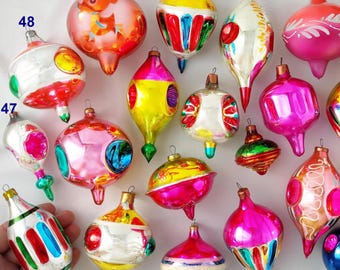 Puede incluir: Una colección de adornos navideños de vidrio vintage en varias formas y colores vibrantes, incluyendo rosa, amarillo, azul, rojo y plata. Los adornos presentan acabados reflectantes, detalles pintados y tapas metálicas, algunos etiquetados con los números del 47 al 66.
