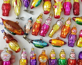 May include: A collection of vintage glass Christmas ornaments featuring various cosmonauts, space-themed figures, and rockets. Many ornaments are marked with the text CCCP. The figures are painted in vibrant colors including red, gold, purple, and green.