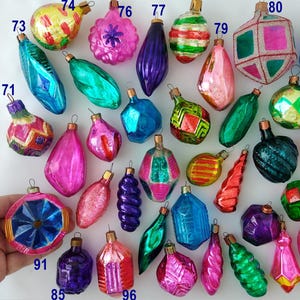 May include: A collection of vintage glass Christmas ornaments in various shapes and vibrant colours including pink, blue, green, purple, and yellow. The ornaments feature metallic finishes, geometric patterns, and ribbed textures with numbered labels.