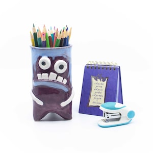 May include: A purple ceramic pencil holder shaped like a monster with white eyes and teeth, filled with coloured pencils. Next to it is a small purple spiral-bound notebook with printed text and a white and teal stapler on a white background.