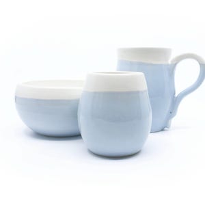 May include: A ceramic set consisting of a bowl, a small rounded cup, and a mug, all featuring a two-tone design with a glossy light blue base and a smooth white upper rim. The mug includes a curved handle on the right side, while the bowl and cup have simple, rounded shapes.