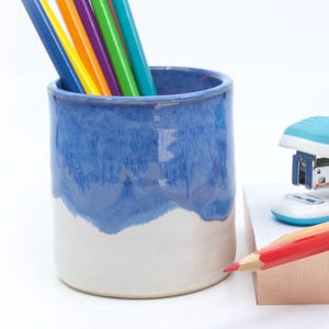 May include: A cylindrical ceramic pencil holder with a two-tone blue and white glaze, containing several coloured pencils. Next to it sits a white and teal stapler, a red pencil, and a stack of light-coloured paper notes on a white background.