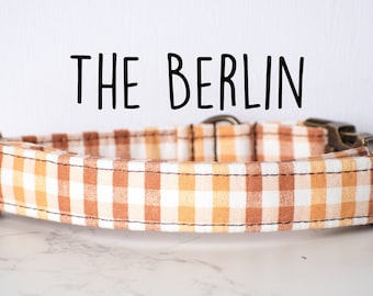May include: A brown and white gingham dog collar with brass hardware. The collar has the text "THE BERLIN" printed above it.