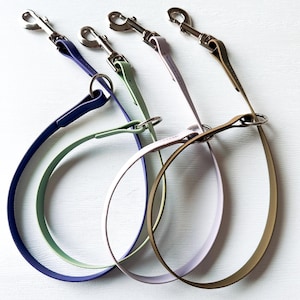 May include: Three dog leashes in different colors: purple, green, and light brown. The leashes are made of a soft, flexible material and have a silver metal clip at the end.