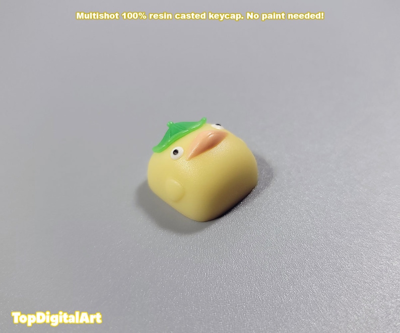 May include: A yellow resin keycap shaped like a cartoon chick wearing a green leaf hat. Multishot 100% resin casted keycap. No paint needed!