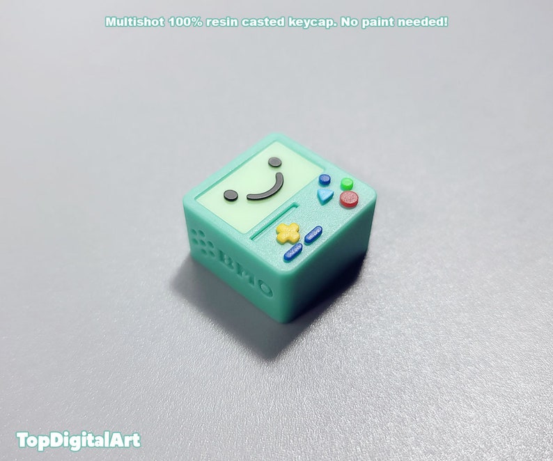 May include: A green and yellow resin keycap shaped like a retro video game console with a smiling face on the screen. The keycap is a multishot design, meaning the colors are layered and not painted.