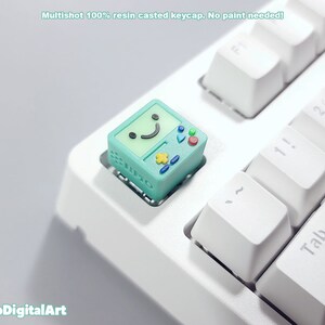 May include: A teal green keycap with a smiling face and buttons, resembling a retro video game console. The keycap is on a white keyboard.