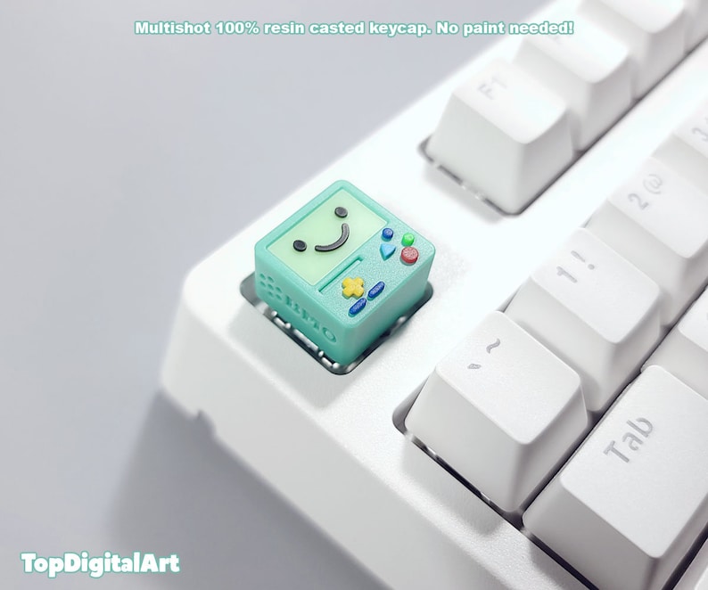 May include: A teal green keycap with a smiling face and buttons, resembling a retro video game console. The keycap is on a white keyboard.