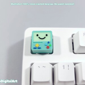 May include: A light blue and green keycap with a smiling face and game controller design. The keycap is made of resin and is designed to fit a standard keyboard.