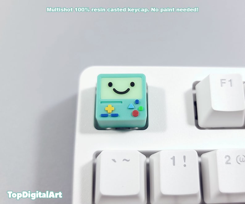 May include: A light blue and green keycap with a smiling face and game controller design. The keycap is made of resin and is designed to fit a standard keyboard.