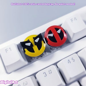 Puede incluir: Dos teclas de resina con los personajes Deadpool y Wolverine. La tecla de Deadpool es roja con detalles negros y la tecla de Wolverine es amarilla con detalles negros. Ambas teclas están en un teclado blanco.
