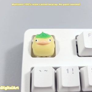 May include: A yellow resin keycap with a green leaf hat. The keycap is shaped like a cartoon duck with a small beak and eyes. The keycap is on a white keyboard.