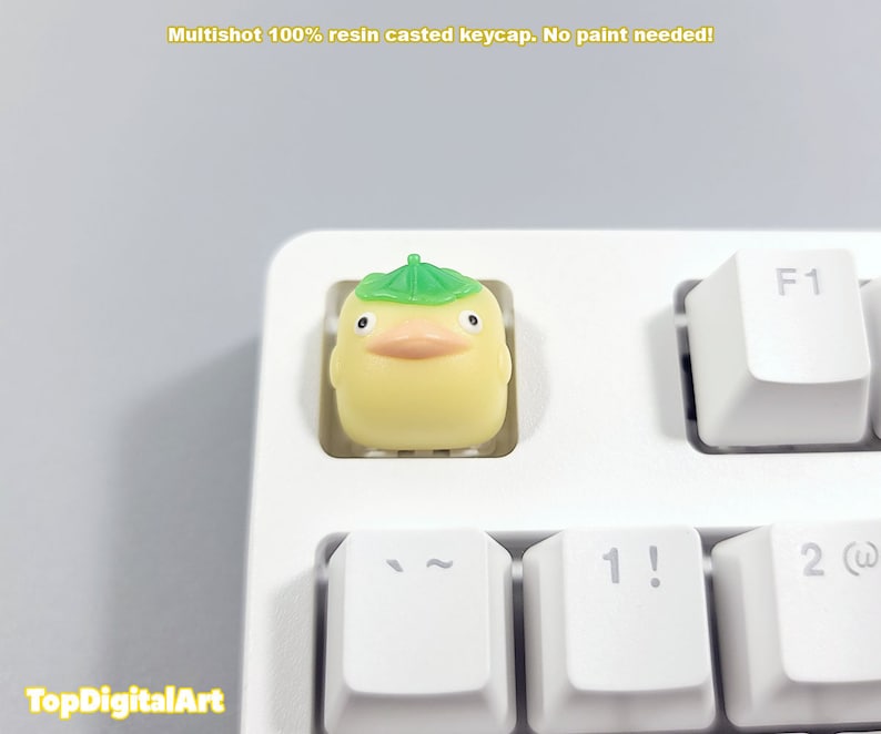 May include: A yellow resin keycap with a green leaf hat. The keycap is shaped like a cartoon duck with a small beak and eyes. The keycap is on a white keyboard.