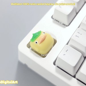 May include: A yellow resin keycap with a green mushroom-shaped hat, designed to look like a cartoon character. The keycap is on a white keyboard.