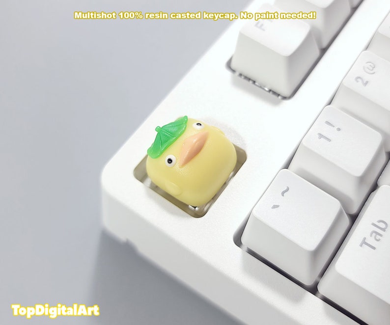 May include: A yellow resin keycap with a green mushroom-shaped hat, designed to look like a cartoon character. The keycap is on a white keyboard.