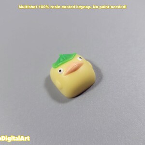 May include: A yellow resin keycap shaped like a duck with a green leaf hat. The keycap is a multishot design, meaning the colors are layered and not painted.
