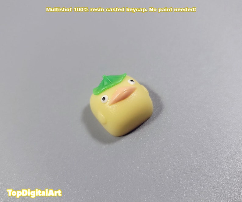 May include: A yellow resin keycap shaped like a duck with a green leaf hat. The keycap is a multishot design, meaning the colors are layered and not painted.