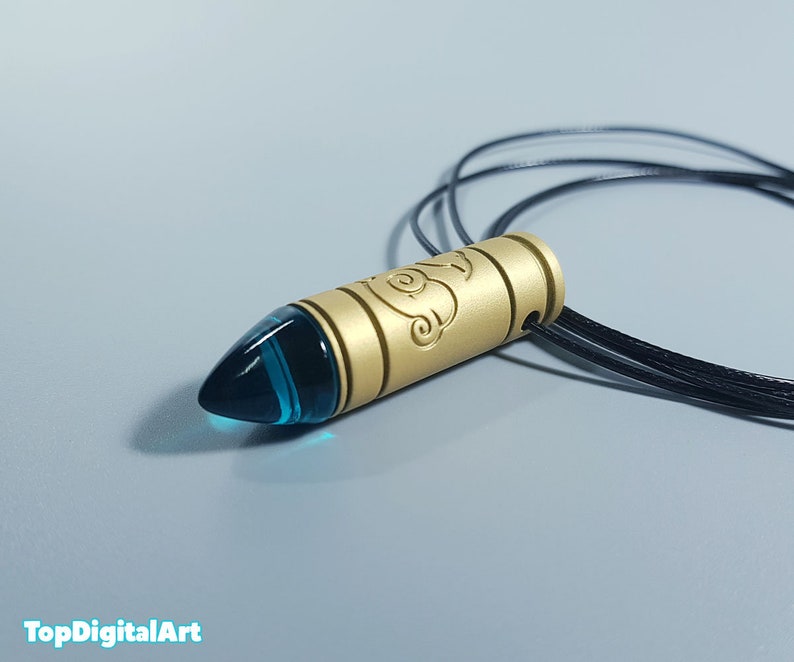 May include: A gold-colored pendant necklace with a blue glass bullet-shaped charm. The charm has a decorative design on the side.
