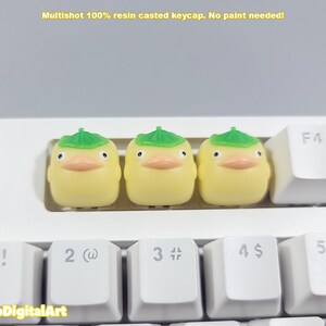 May include: Three yellow duck keycaps with green leaf hats. The keycaps are on a white keyboard with black keys.
