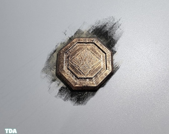 May include: A bronze-colored, octagonal medallion with a detailed central design. The medallion has a raised border and a textured surface. The background is a light gray with a dark, textured brushstroke.