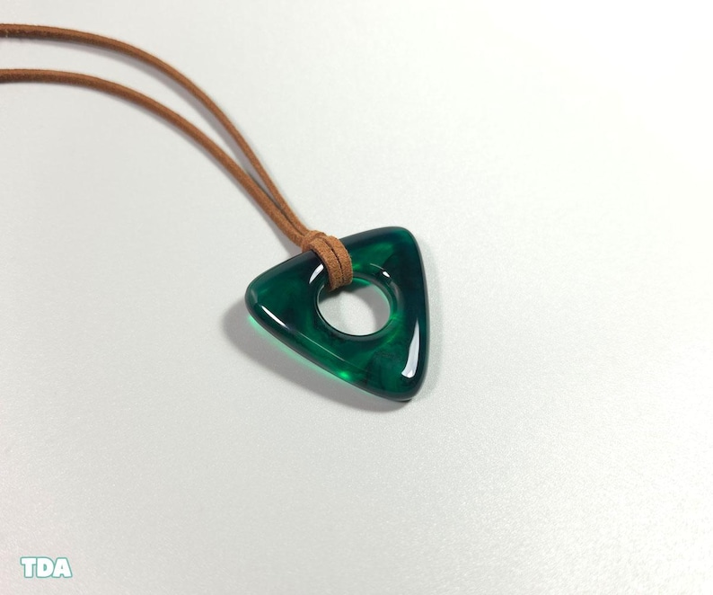 May include: A triangular pendant necklace with a hole in the center. The pendant is a deep emerald green color and is attached to a brown leather cord. The necklace is displayed on a white surface.