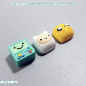 May include: Three resin keycaps featuring characters from the animated series Adventure Time. The keycaps are green, white, and yellow. The green keycap features a smiling face with a star and a triangle. The white keycap features a smiling face with a white hat. The yellow keycap features a smiling face with black eyes and a black nose.