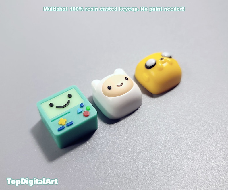May include: Three resin keycaps featuring characters from the animated series Adventure Time. The keycaps are green, white, and yellow. The green keycap features a smiling face with a star and a triangle. The white keycap features a smiling face with a white hat. The yellow keycap features a smiling face with black eyes and a black nose.