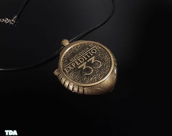 May include: A bronze-colored pendant with the text "CLAIR OBSCUR EXPEDITION 33" on a black cord. The pendant has a circular design with a textured surface and a detailed border. The background is black.