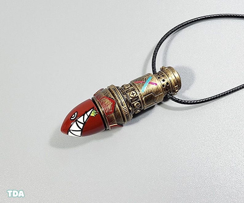 May include: A gold and red necklace with a detailed, textured pendant shaped like a bullet with a toothy, cartoonish face. The pendant has a black cord.