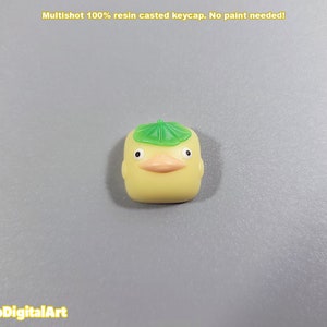 May include: A yellow resin keycap shaped like a duck wearing a green leaf hat. The keycap is a multishot design, meaning the colors are layered and not painted.