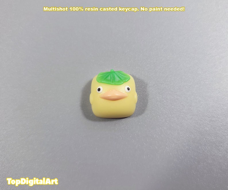 May include: A yellow resin keycap shaped like a duck wearing a green leaf hat. The keycap is a multishot design, meaning the colors are layered and not painted.