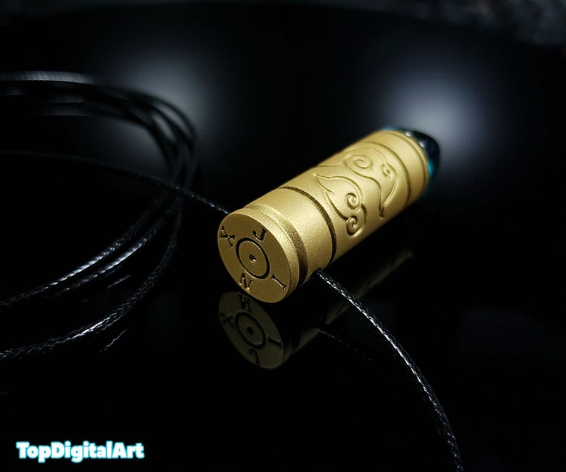 May include: A gold-colored bullet casing necklace with a decorative design. The casing has a black cord attached to it.