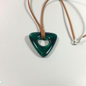 May include: A triangular, emerald green pendant with a circular hole, suspended on a brown leather cord necklace. The pendant has a glossy finish and is attached to a silver clasp. The necklace is displayed on a white surface.