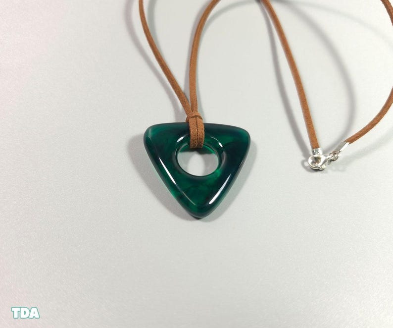 May include: A triangular, emerald green pendant with a circular hole, suspended on a brown leather cord necklace. The pendant has a glossy finish and is attached to a silver clasp. The necklace is displayed on a white surface.