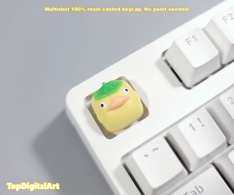 May include: A yellow resin keycap with a green leaf hat, shaped like a duck, on a white keyboard. The keycap is a multishot 100% resin casted keycap. No paint needed!