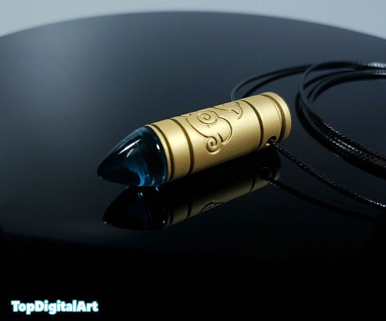 May include: A gold bullet-shaped pendant with a blue glass tip, hanging from a black cord. The pendant has an intricate design on the side.