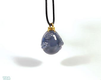 May include: A blue-grey pendant necklace with a gold-colored bail and a black cord. The pendant has a rounded shape with a textured surface. The necklace is displayed against a white background.