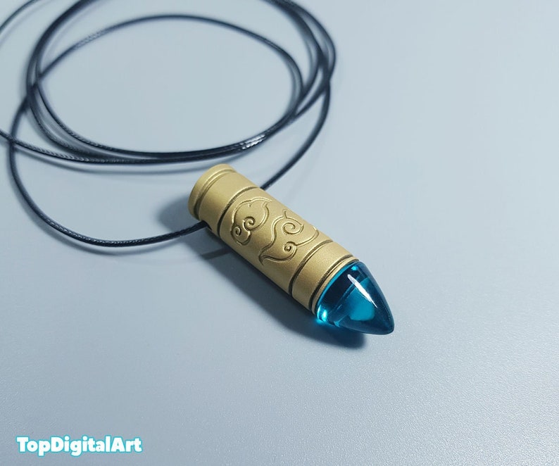 May include: A gold-colored pendant necklace with a blue glass tip. The pendant is shaped like a bullet and has a design of clouds on it.