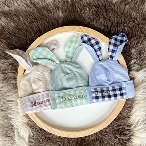 May include: Three ribbed knit baby hats with bunny ears, each with a name embroidered on the front. The hats are beige, mint green, and light blue, with gingham ear linings in coordinating colors. The names embroidered are "Mason", "Damien", and "William".