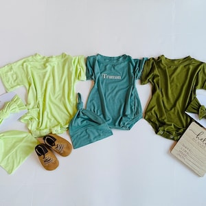 May include: A collection of baby clothing and accessories in shades of green and yellow. Includes a lime green onesie, a teal romper with the name "Truman", and an olive green onesie. Accessories include matching headbands and hats, and a pair of brown leather baby shoes.