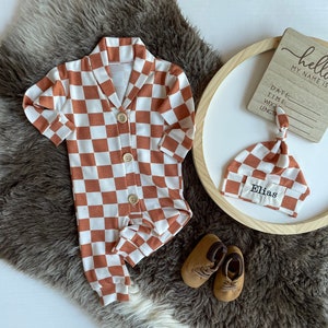 May include: A brown and white checkered baby onesie with a button closure. A matching brown and white checkered hat with the name "Elias" embroidered on it. Brown leather baby shoes. A wooden sign with the text "hello my name is" and blank lines for information.