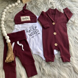 May include: A baby outfit set in burgundy with a white onesie that says "hello world" with an arrow, burgundy pants with a brown tassel, a burgundy cardigan with wooden buttons, and a burgundy knit hat with the name "Seth" embroidered on it.