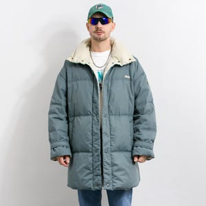 Può includere: Un lungo piumino verde acqua con colletto color crema. Il cappotto è abbottonato e indossato con jeans blu, una maglietta bianca e un berretto da baseball verde. La persona indossa anche occhiali da sole e scarpe da ginnastica.