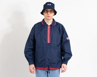 May include: A navy blue windbreaker jacket with a red zipper and red trim. The jacket has a small American flag patch on the right sleeve and a drawstring at the waist. The model is wearing a blue bucket hat with the word "FILA" in white.