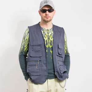 Puede incluir: Un chaleco azul pizarra con múltiples bolsillos y cremalleras. El chaleco se lleva sobre una camisa de manga larga con un estampado verde y amarillo. La persona lleva una gorra de béisbol gris y gafas de sol.